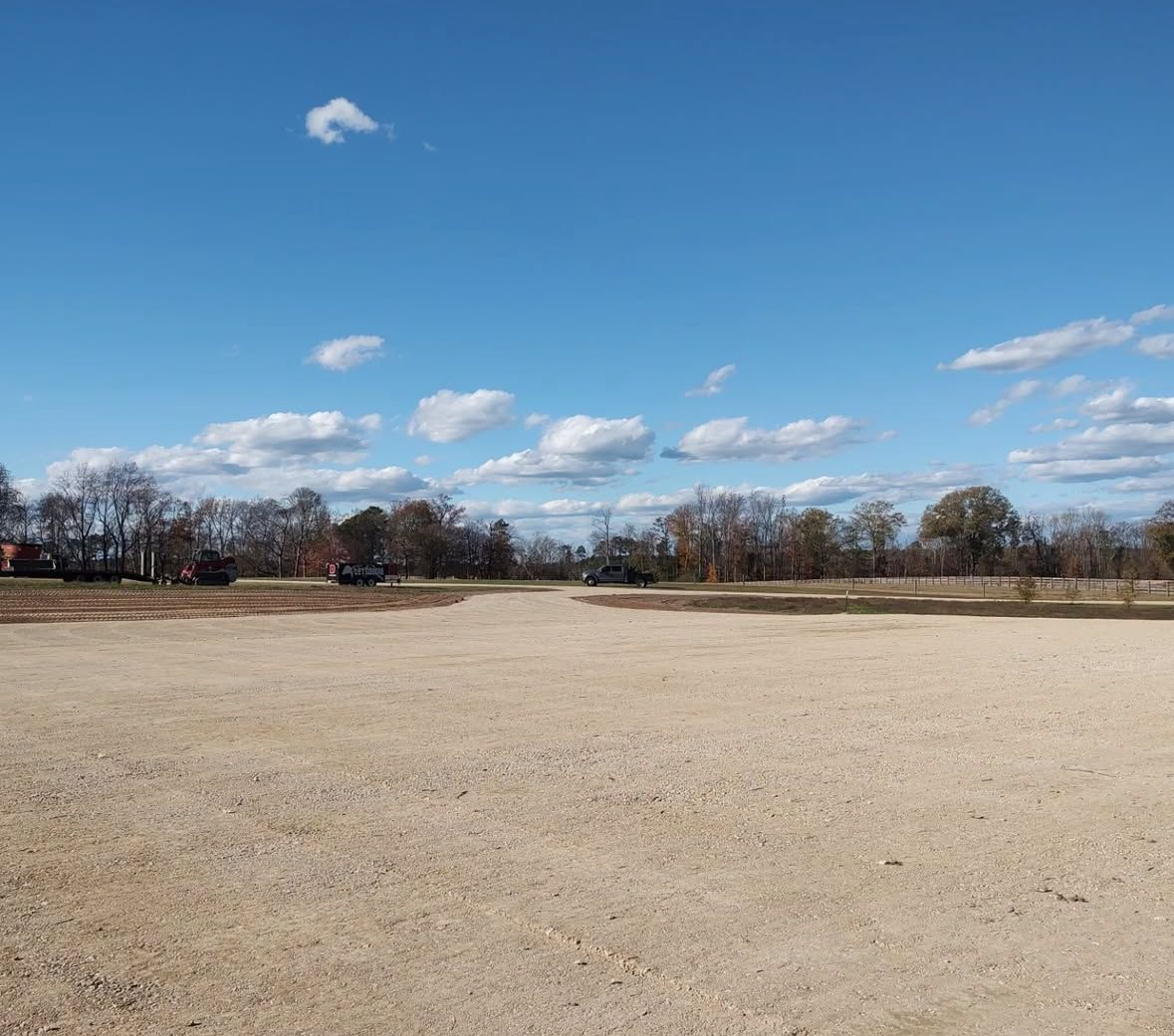 Open field with gravel surface, trees and buildings in the background under a blue sky with scattered clouds.
