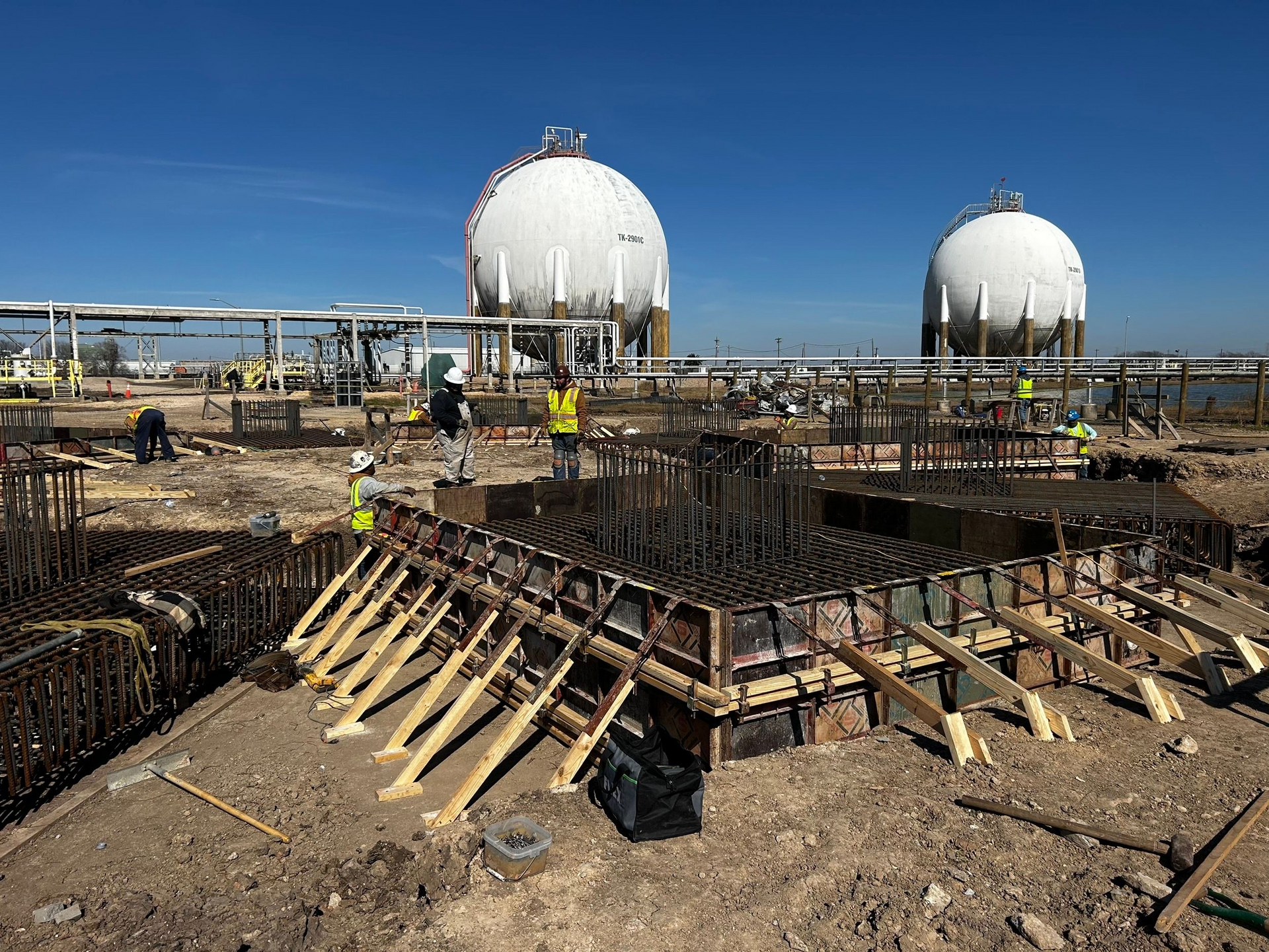 Construction site with workers, rebar, wooden forms, and two white spherical tanks against a blue sky.