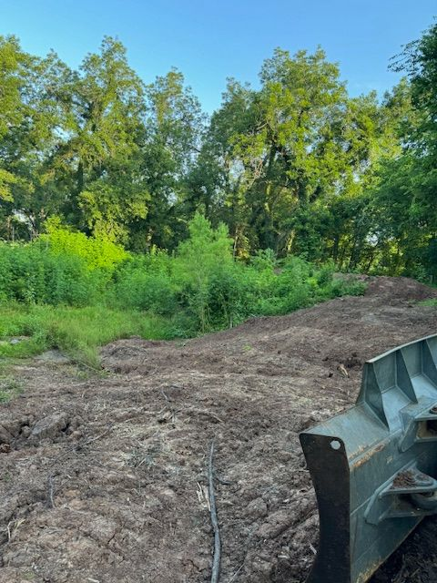 Dirt pile with greenery, trees in background, and a piece of machinery in the foreground under a blue sky.