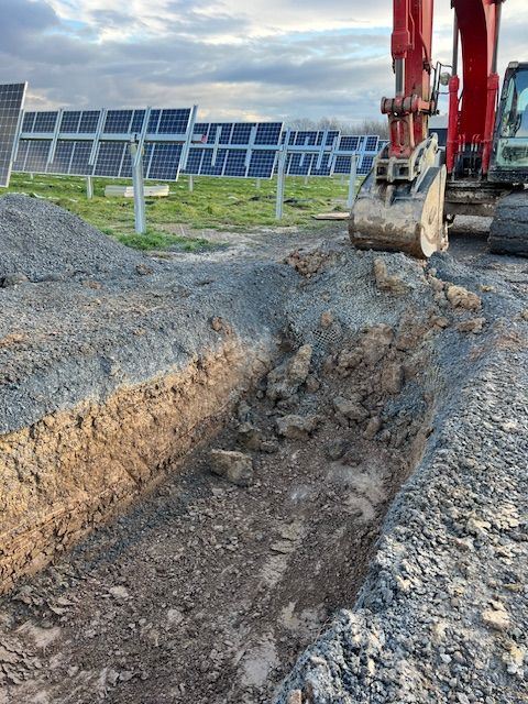 An excavator digs a trench in rocky soil, solar panels in the background.