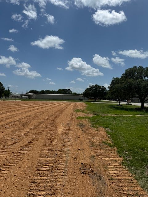 Brown soil with tractor tracks, green grass, trees, buildings, and a cloudy blue sky.