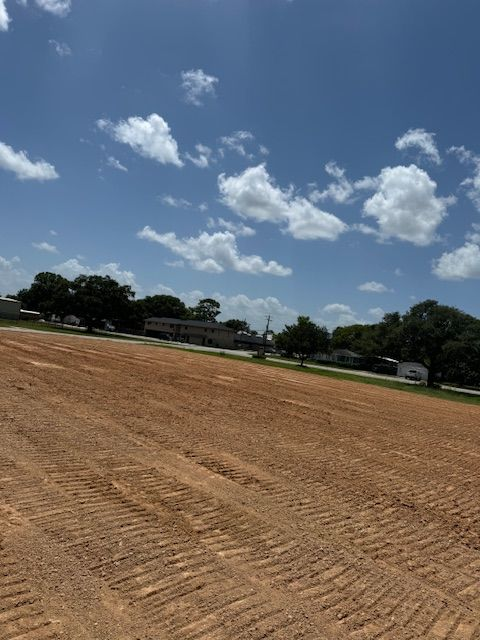 Cleared brown field with tire tracks, trees, buildings, and a blue sky with clouds.