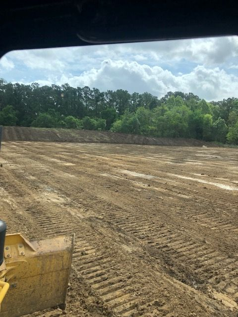 Bulldozer blade in foreground, leveling dirt on a cleared construction site; trees and cloudy sky in background.