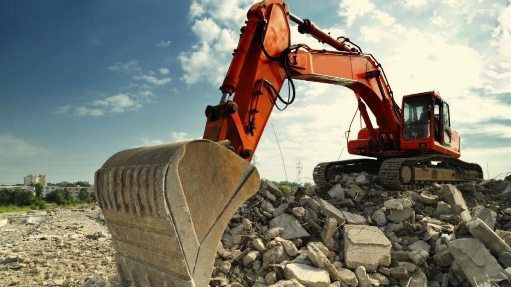 Orange excavator on a pile of rubble against a blue sky with clouds.