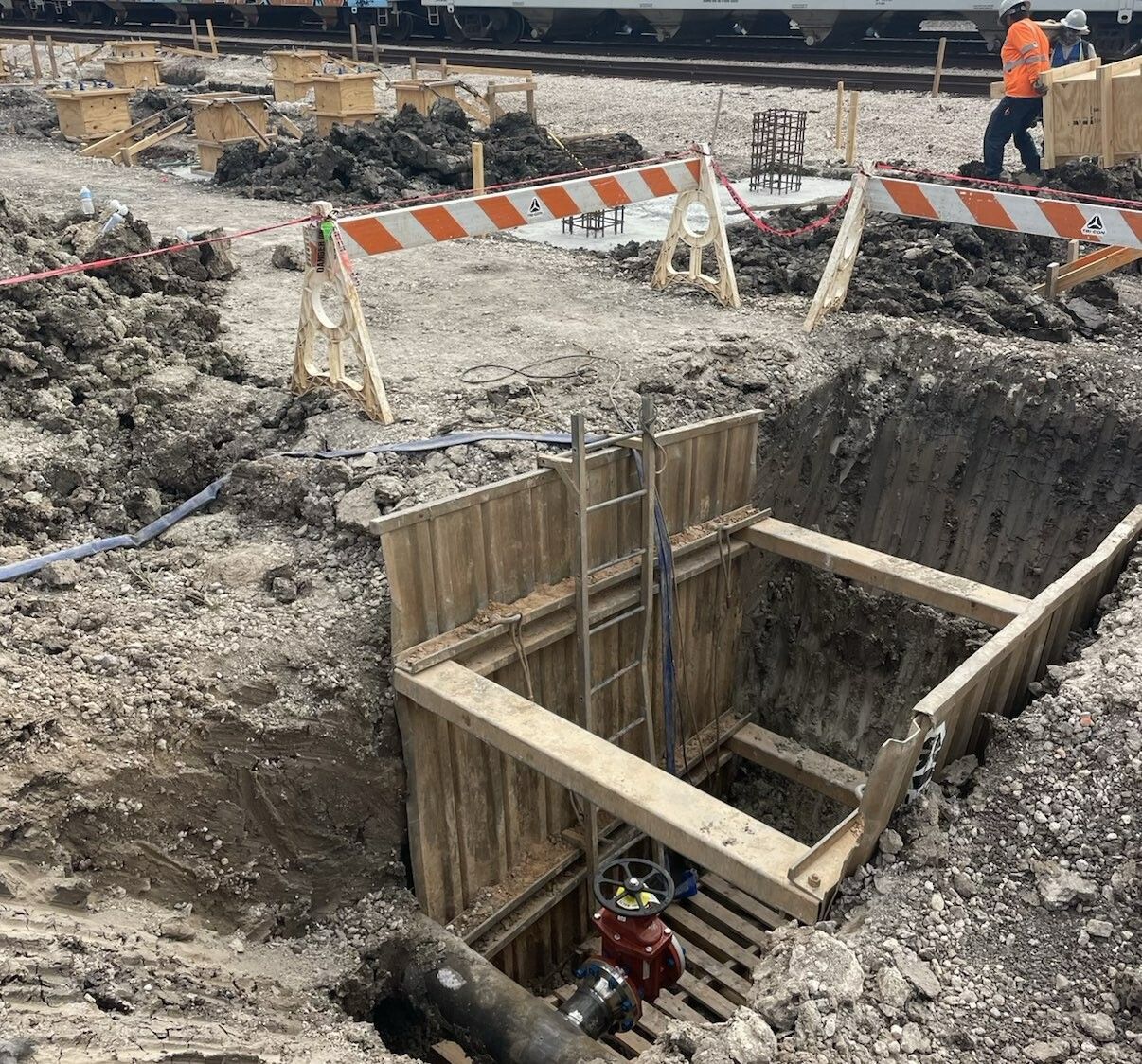 Construction site: Excavation with wooden shoring, ladder. Worker in orange vest, train in background.