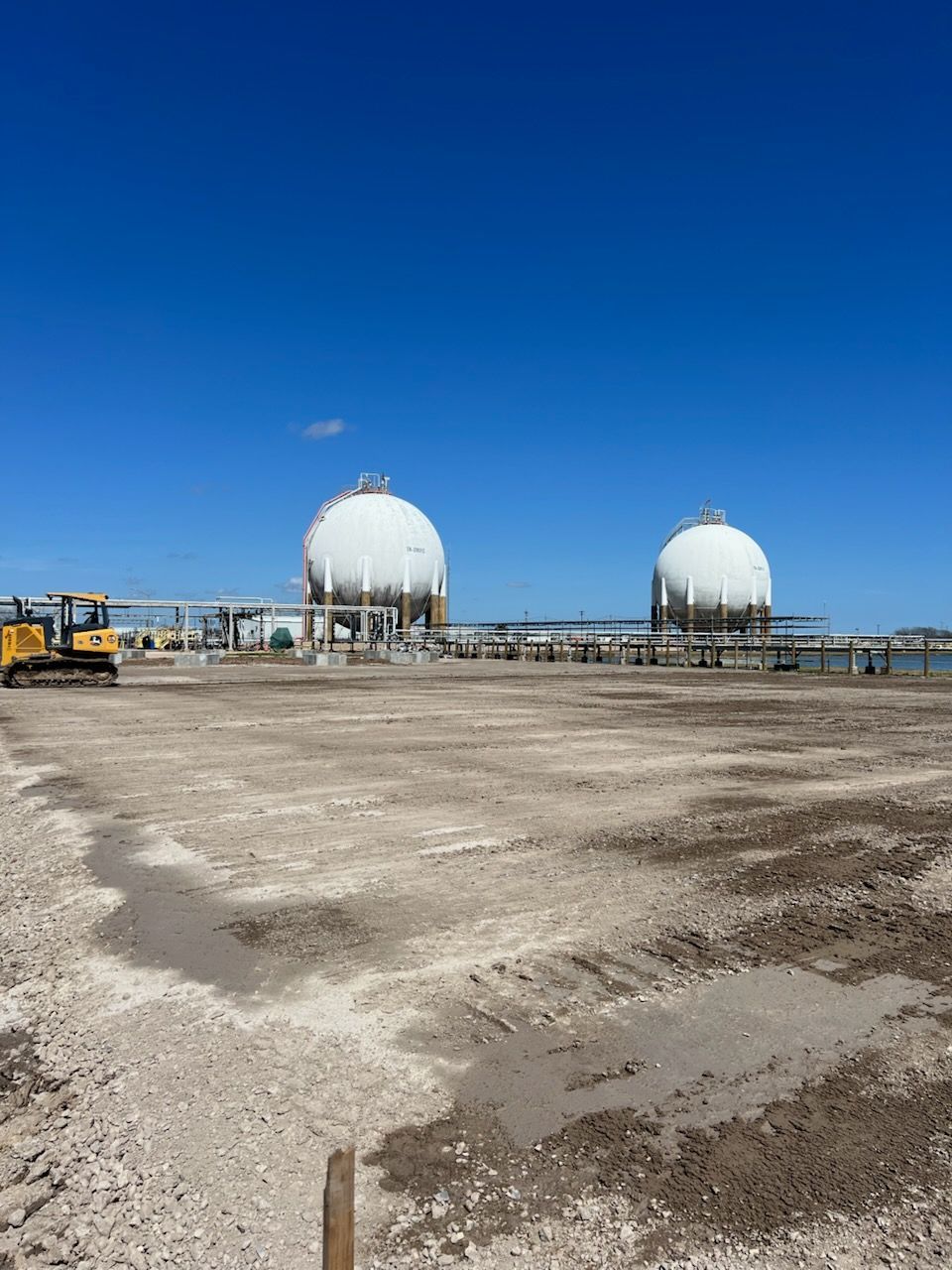 Two white spherical storage tanks on a dirt lot under a bright blue sky. A yellow construction vehicle is visible.