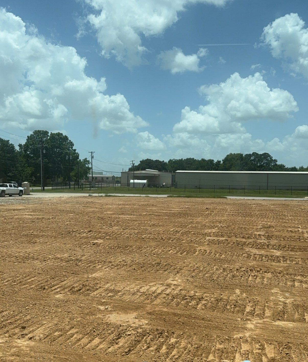 Brown dirt field under a bright blue sky with puffy clouds; a building and trees are in the background.