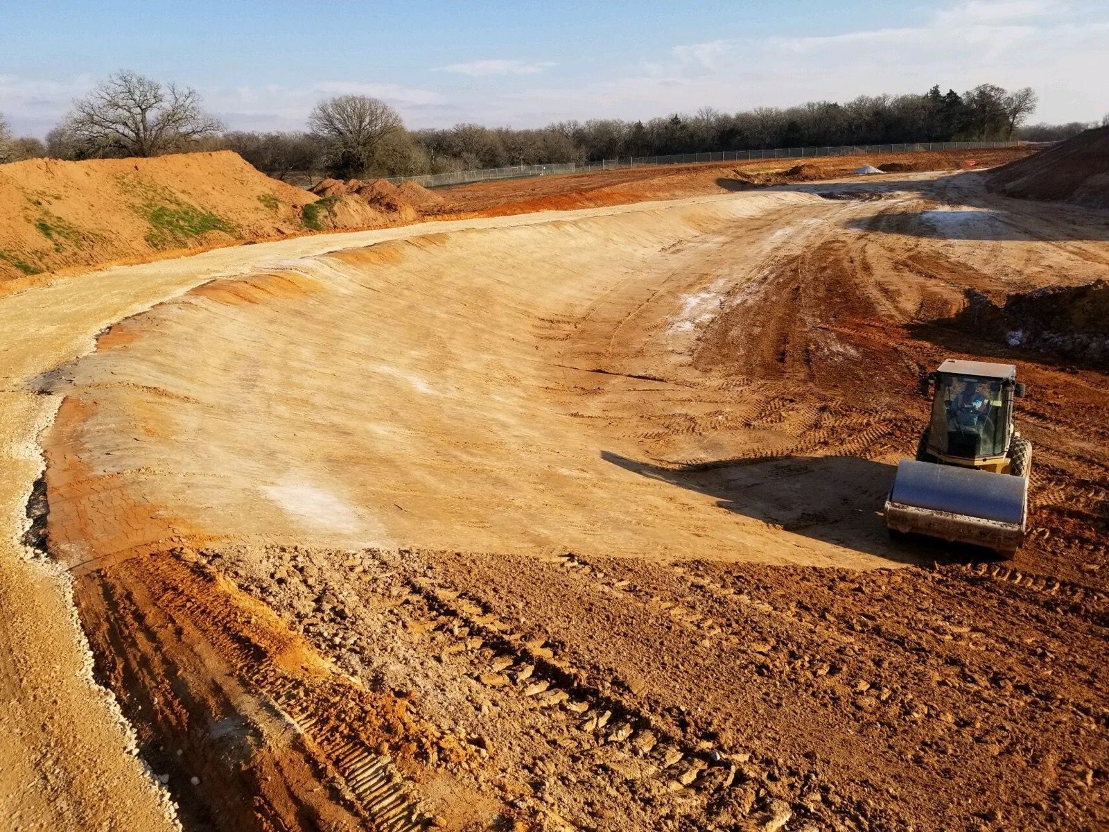Brown, sandy ground with tractor tracks, houses in background under a sunny sky.
