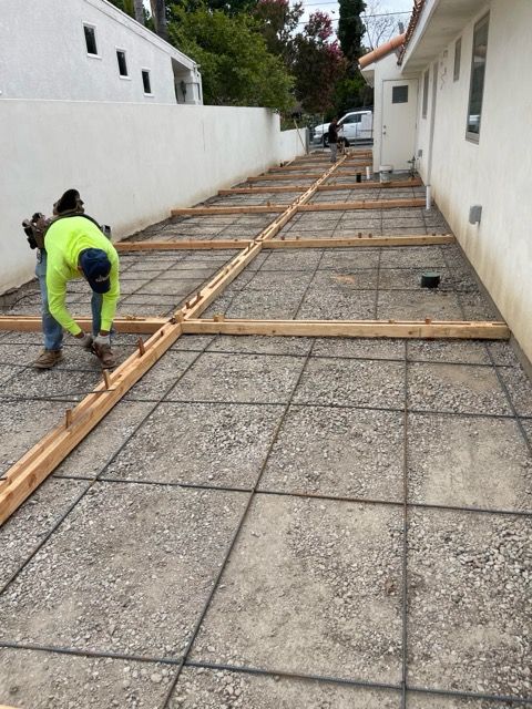 Construction worker laying rebar grid for concrete, wooden forms define sections along a wall and gravel.