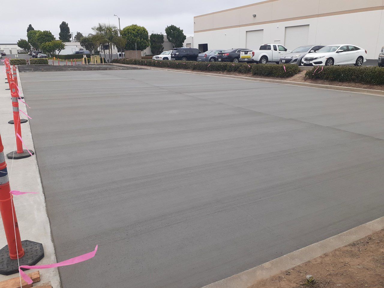 Newly poured concrete parking lot with orange safety markers. Cars parked along the edge in the background.