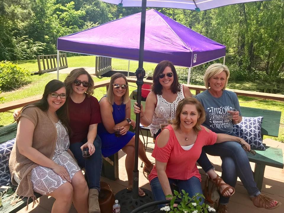 A group of women smiles on a patio under a purple umbrella and canopy.