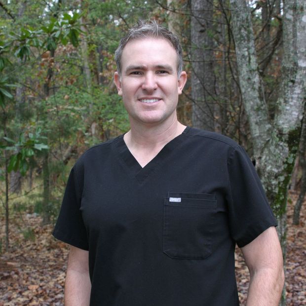 Smiling person in a black shirt standing outdoors among trees and foliage