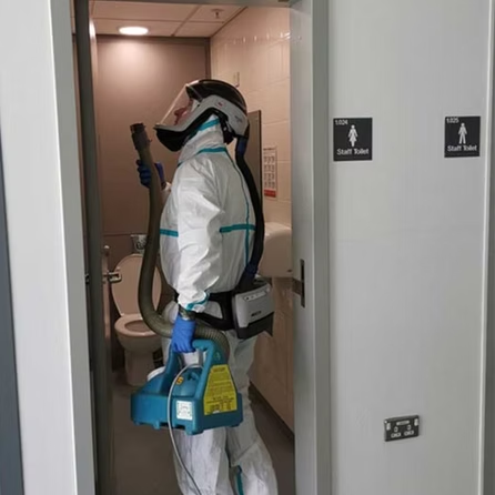 A man in a protective suit is standing in a bathroom holding a vacuum cleaner.