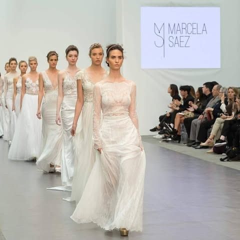 Modelos con vestidos blancos desfilan por una pasarela en un desfile de moda; público al fondo.