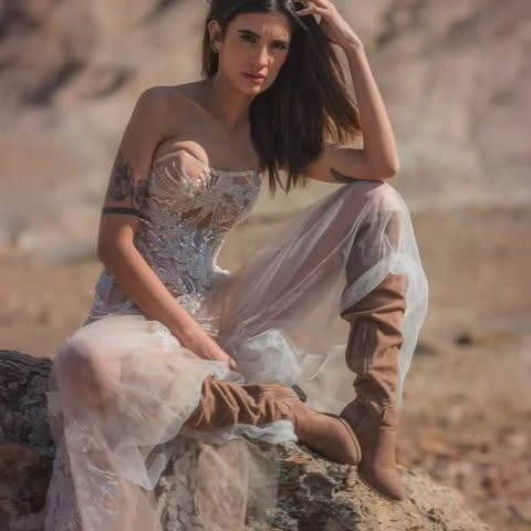 Mujer con vestido blanco vaporoso y botas sentada sobre una roca, con el brazo levantado