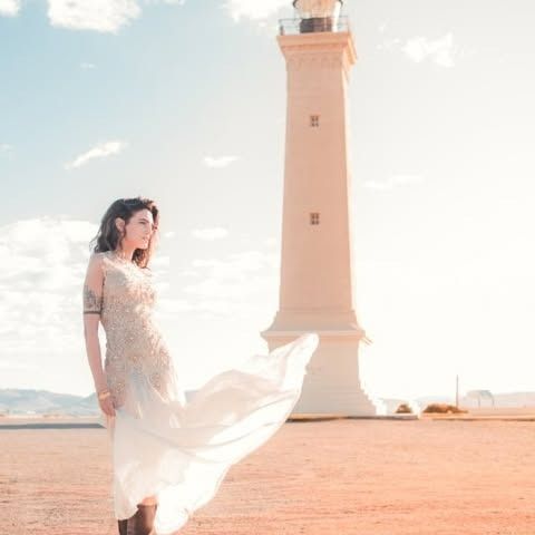 Una mujer con un vaporoso vestido blanco está de pie cerca de un alto faro blanco en un día soleado.
