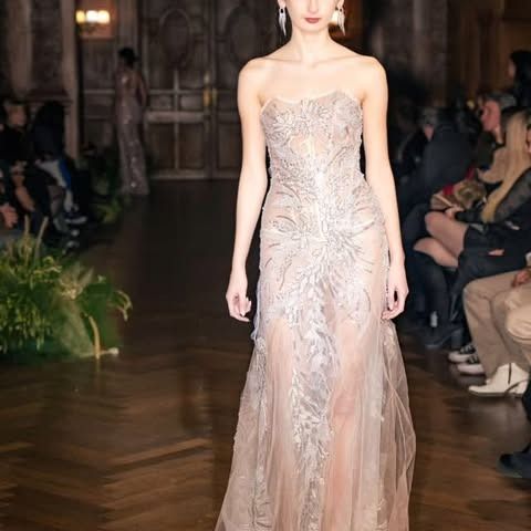 Una modelo con un vestido sin tirantes desfila por la pasarela. El vestido está adornado con bordados plateados