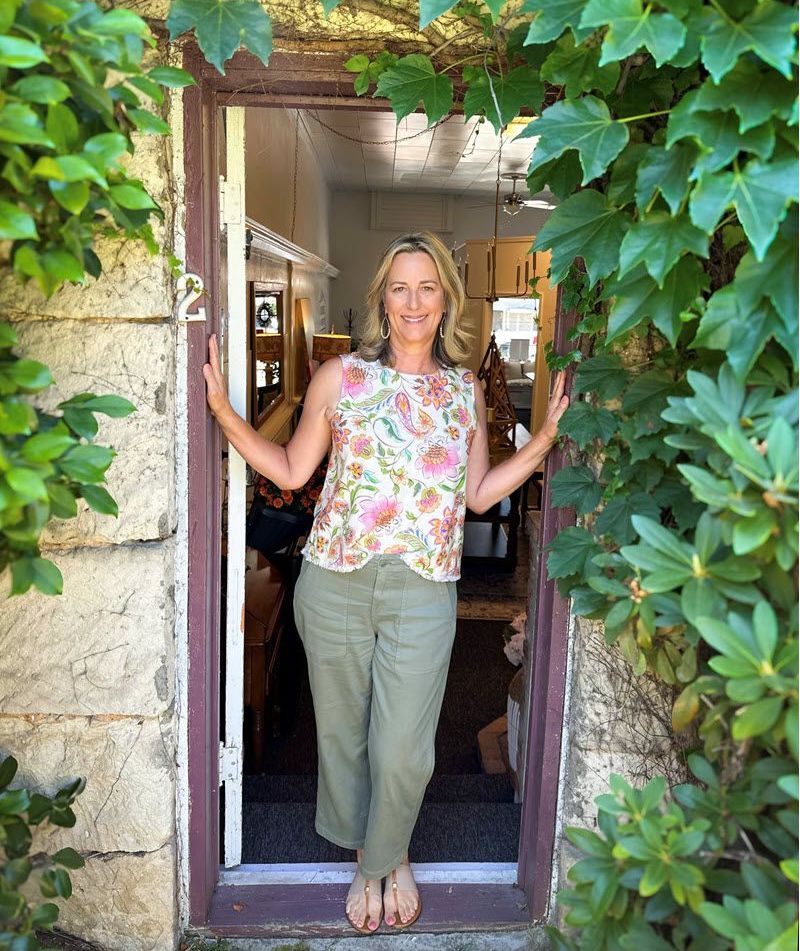 Woman in floral top and green pants stands in doorway, arms outstretched.