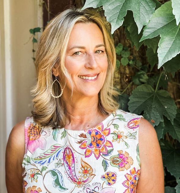 A woman in a floral dress is smiling in front of a tree.