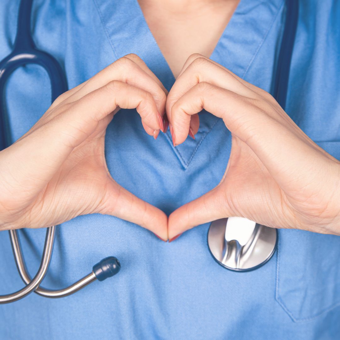 Nurse in blue scrubs forms heart with hands, stethoscope draped around neck.