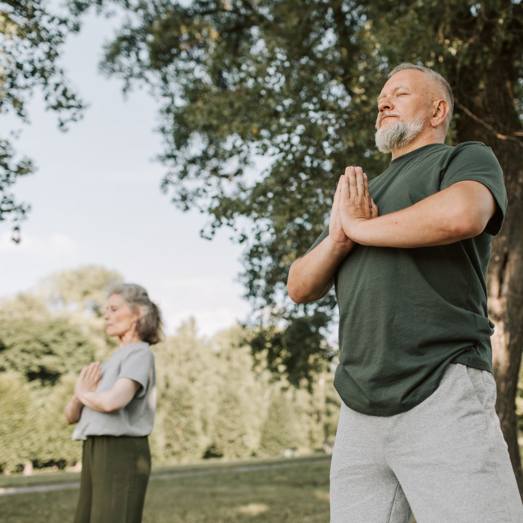 Two people outdoors, hands together in prayer pose, eyes closed, practicing yoga in a park.