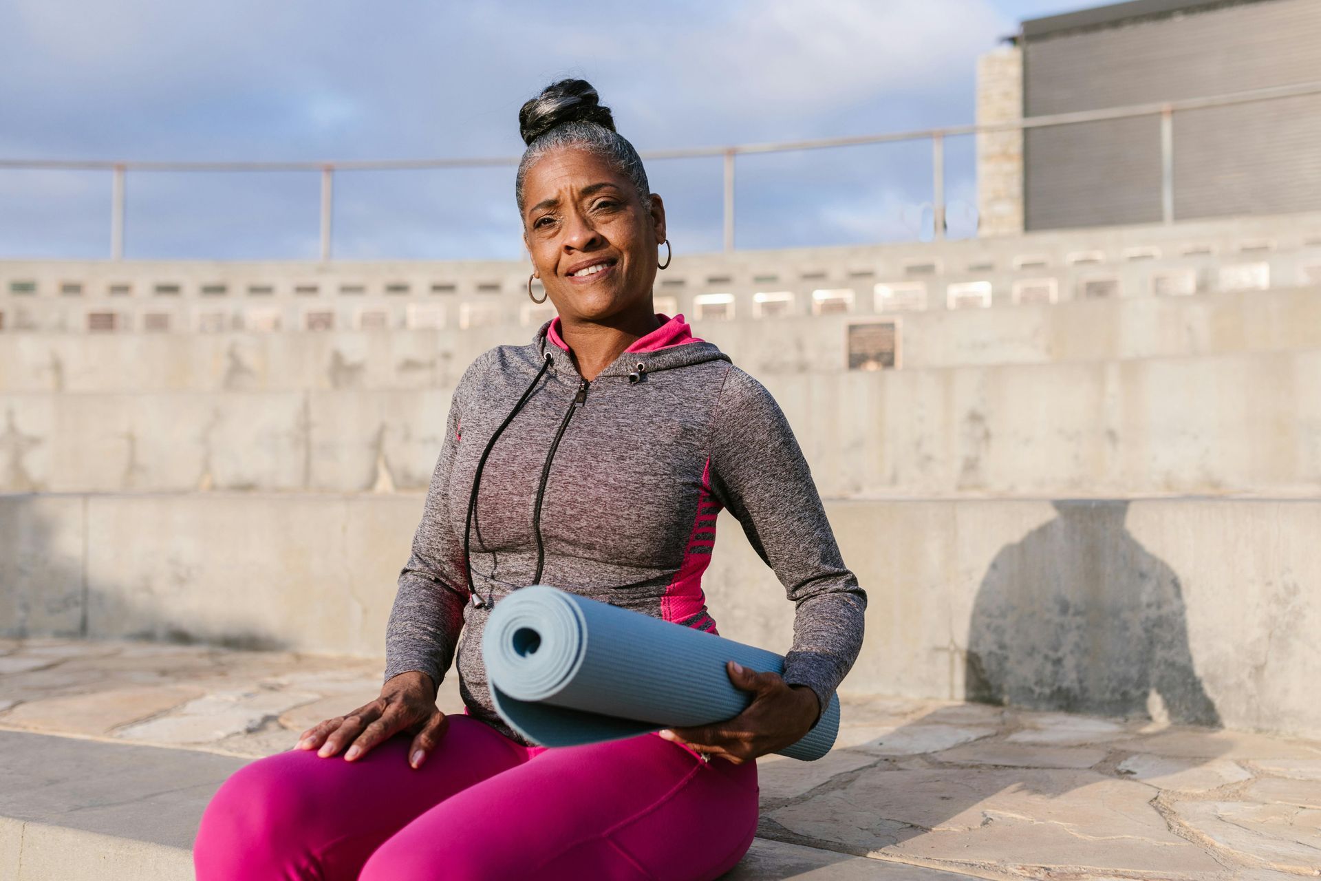 A woman smiles, holding a blue yoga mat in outdoor amphitheater. She wears pink leggings and a gray hoodie.