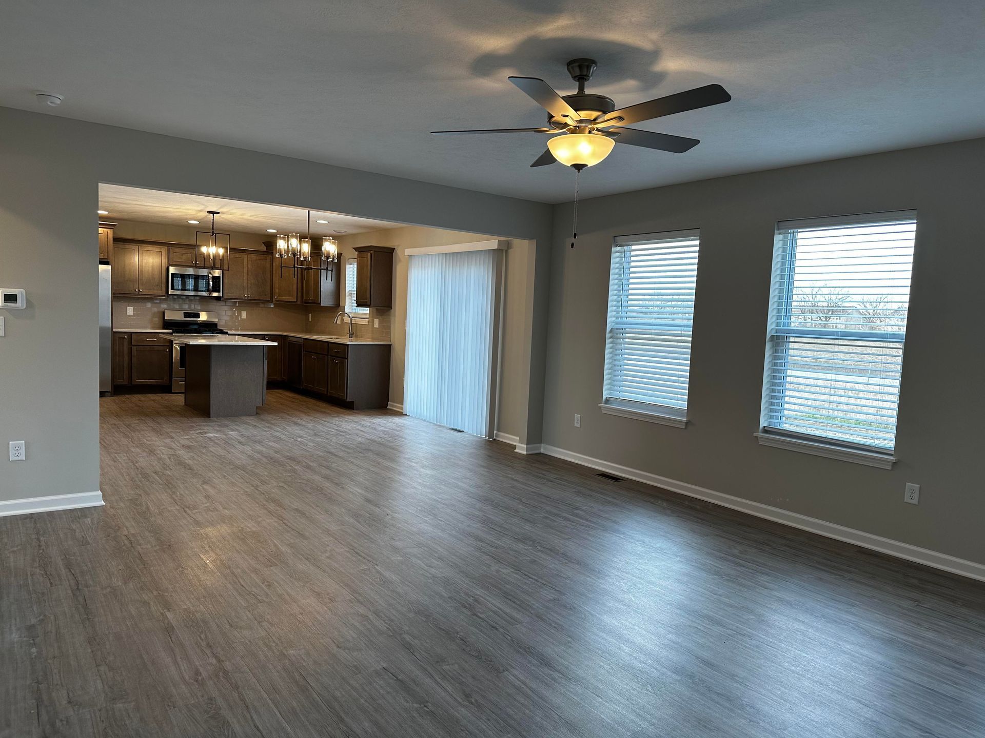 An empty living room with a ceiling fan and a kitchen in the background.