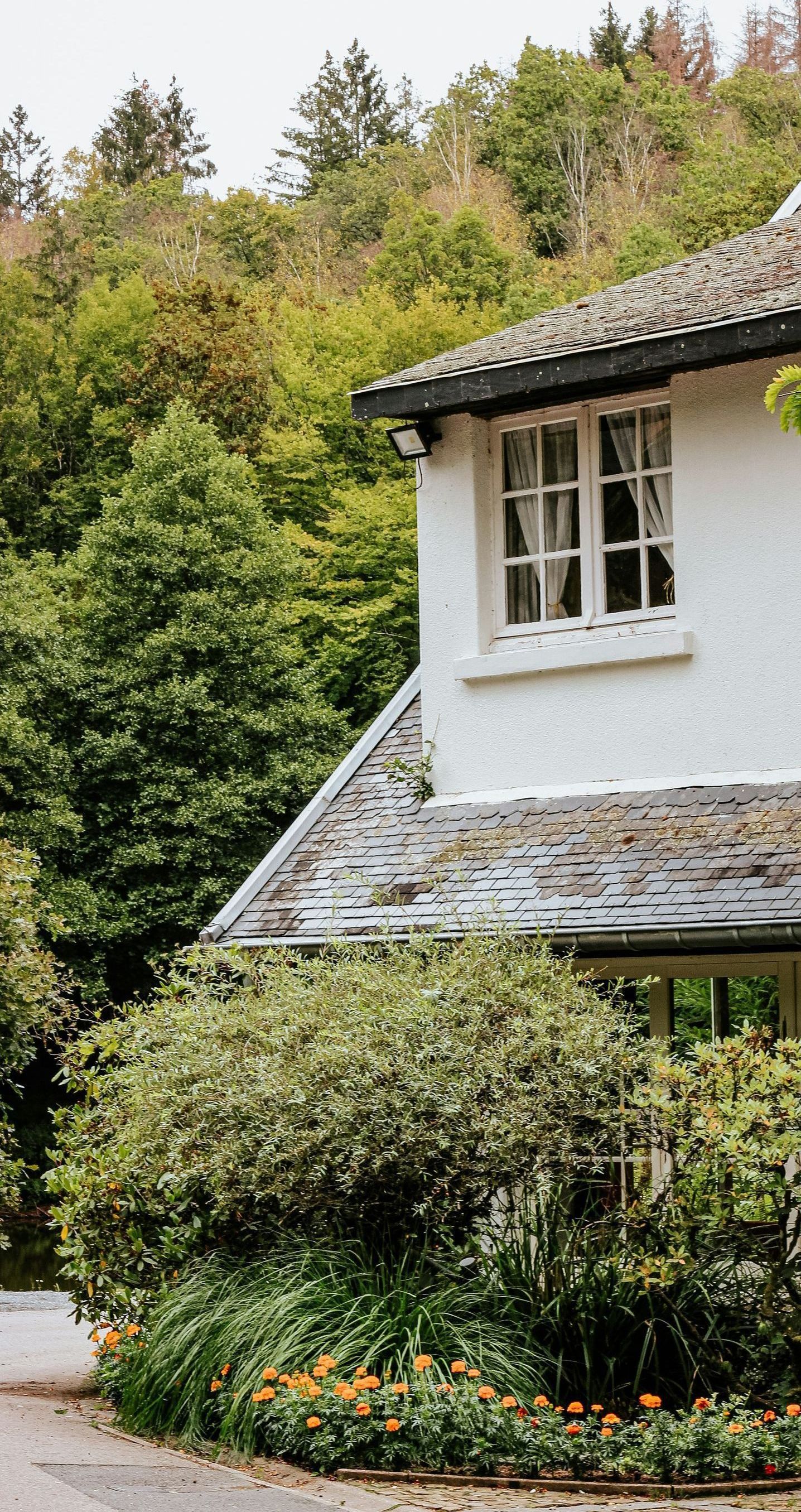 A white house with a stone roof is surrounded by trees and bushes.