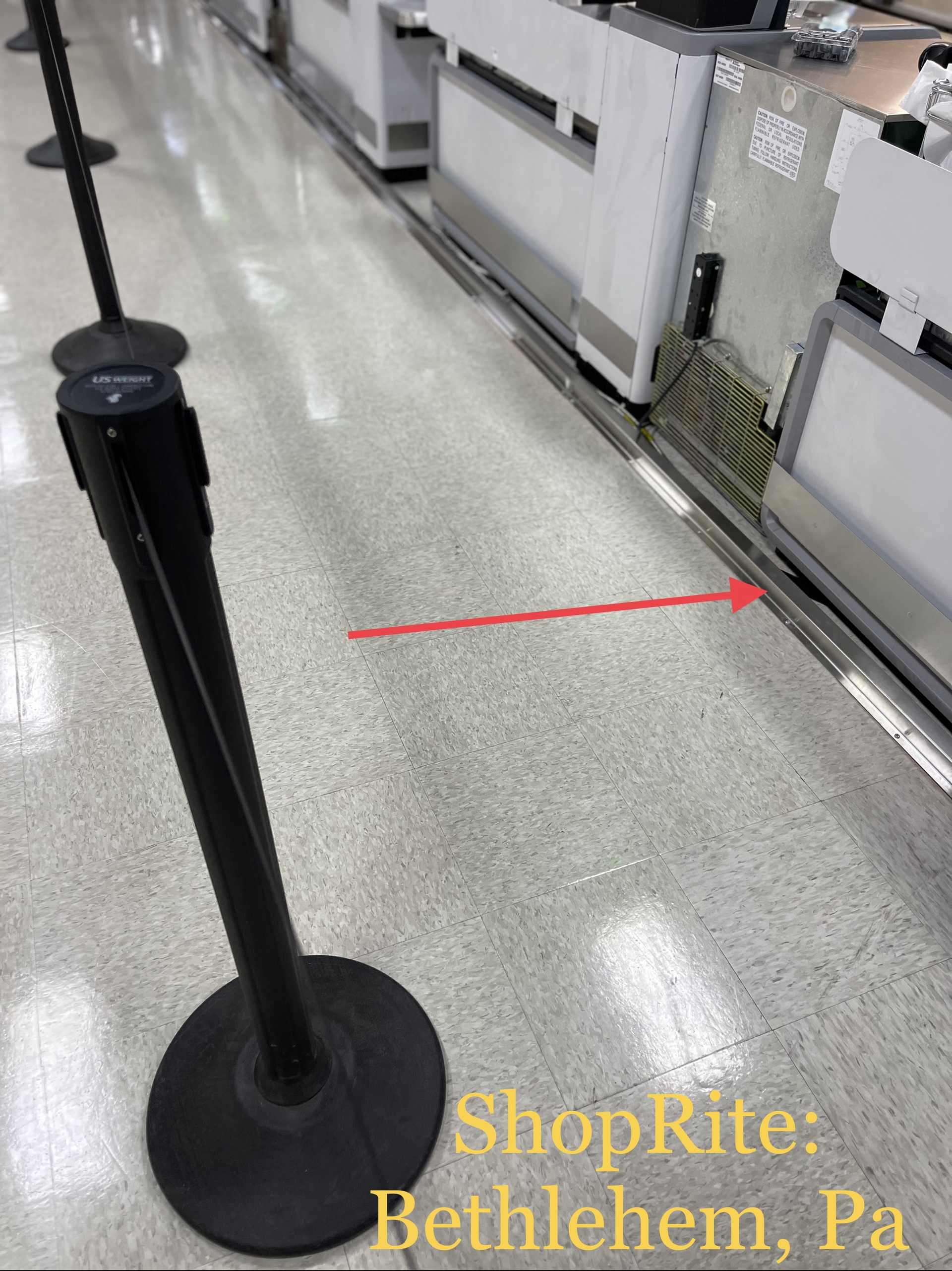 Store aisle with a black stanchion and a red arrow pointing to the floor near shelving.