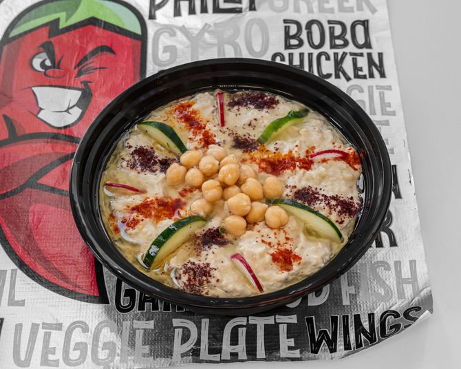 Hummus in black bowl, garnished with chickpeas, cucumber, and paprika, on a patterned paper with restaurant logos.
