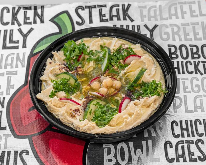 Hummus bowl topped with vegetables, in a black container, on a food-themed background.