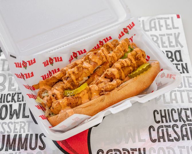 A loaded sub sandwich in a white container, with fried food and sauce, lettuce and pickles.
