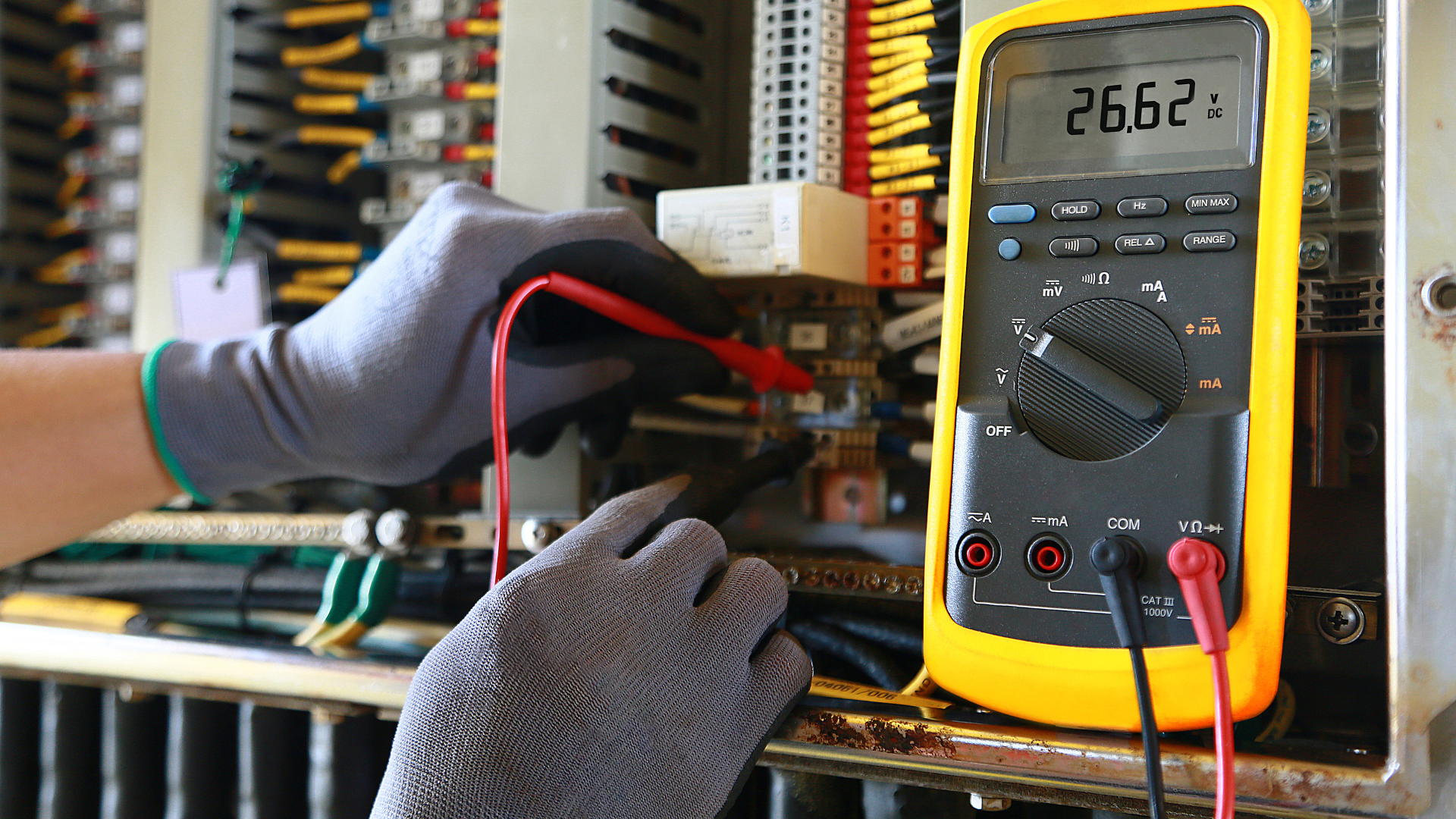 Electrician using a multimeter to measure voltage inside an electrical panel. Gloves and wires are visible.