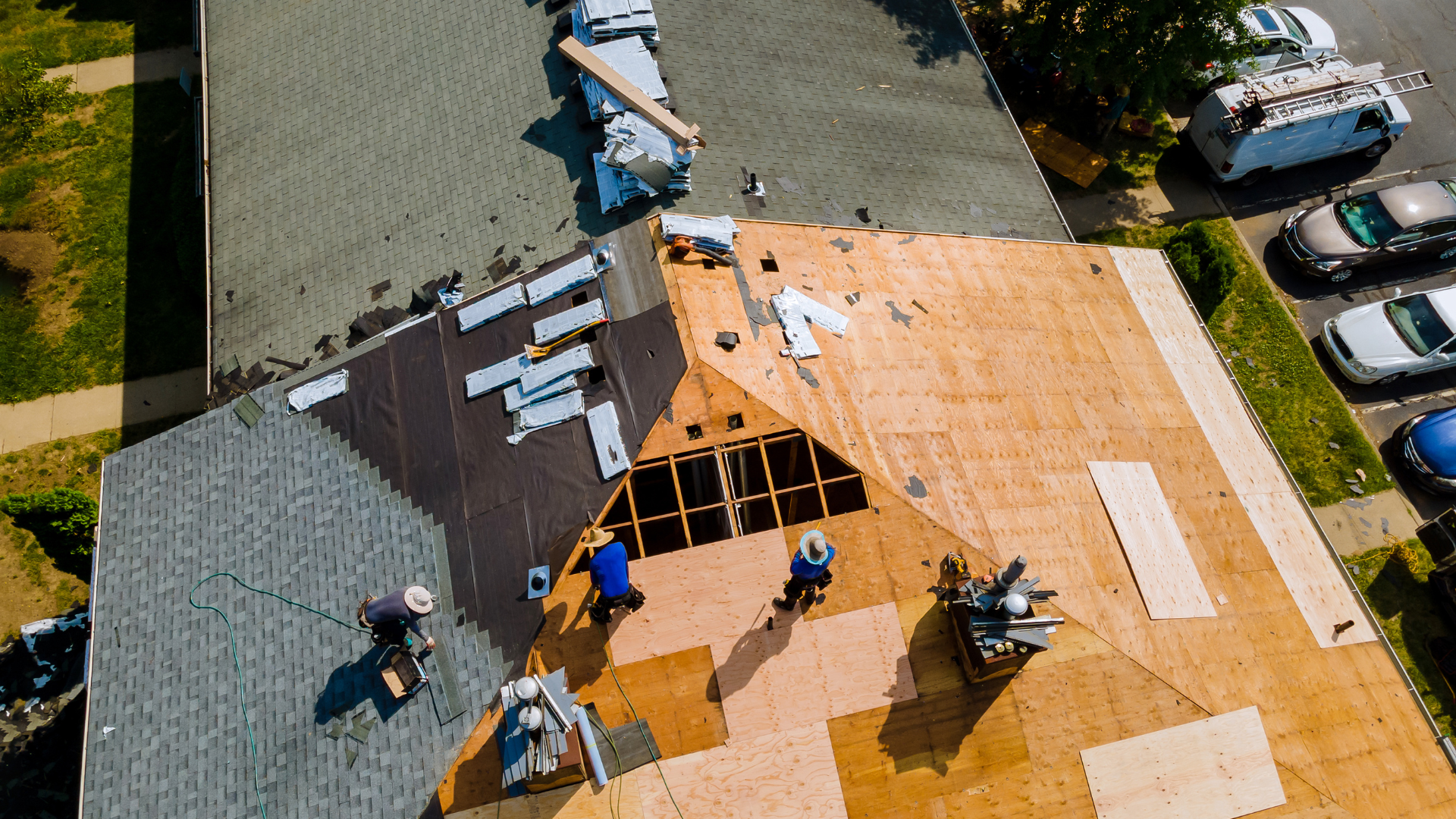 Roofers on a house roof replacing shingles with visible wood support structure. Cars and a service truck parked nearby.
