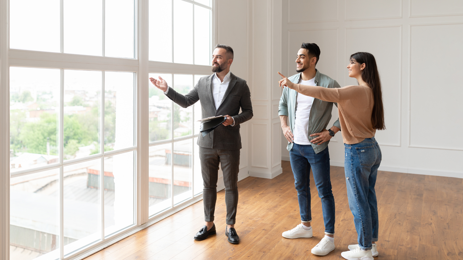 Real estate agent showing large windows to a couple in an empty room; the woman points.