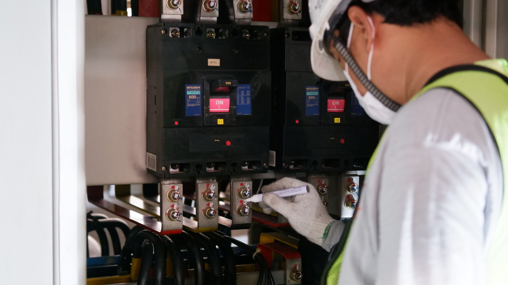 Electrician in safety gear, inspecting electrical panel, marking connections with a pen.