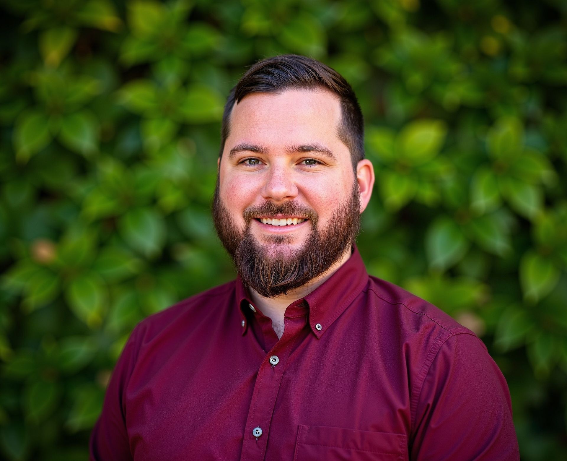 Man with a beard and burgundy shirt smiles in front of green foliage.