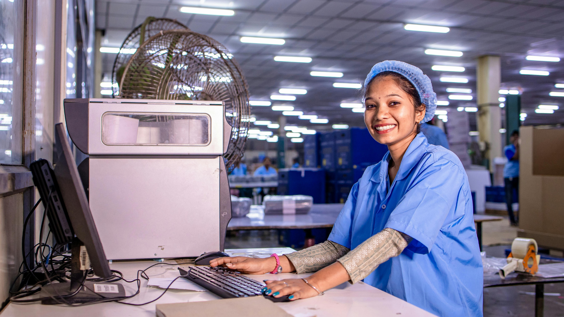 Woman in blue work uniform, smiling, typing at a computer in a factory setting.