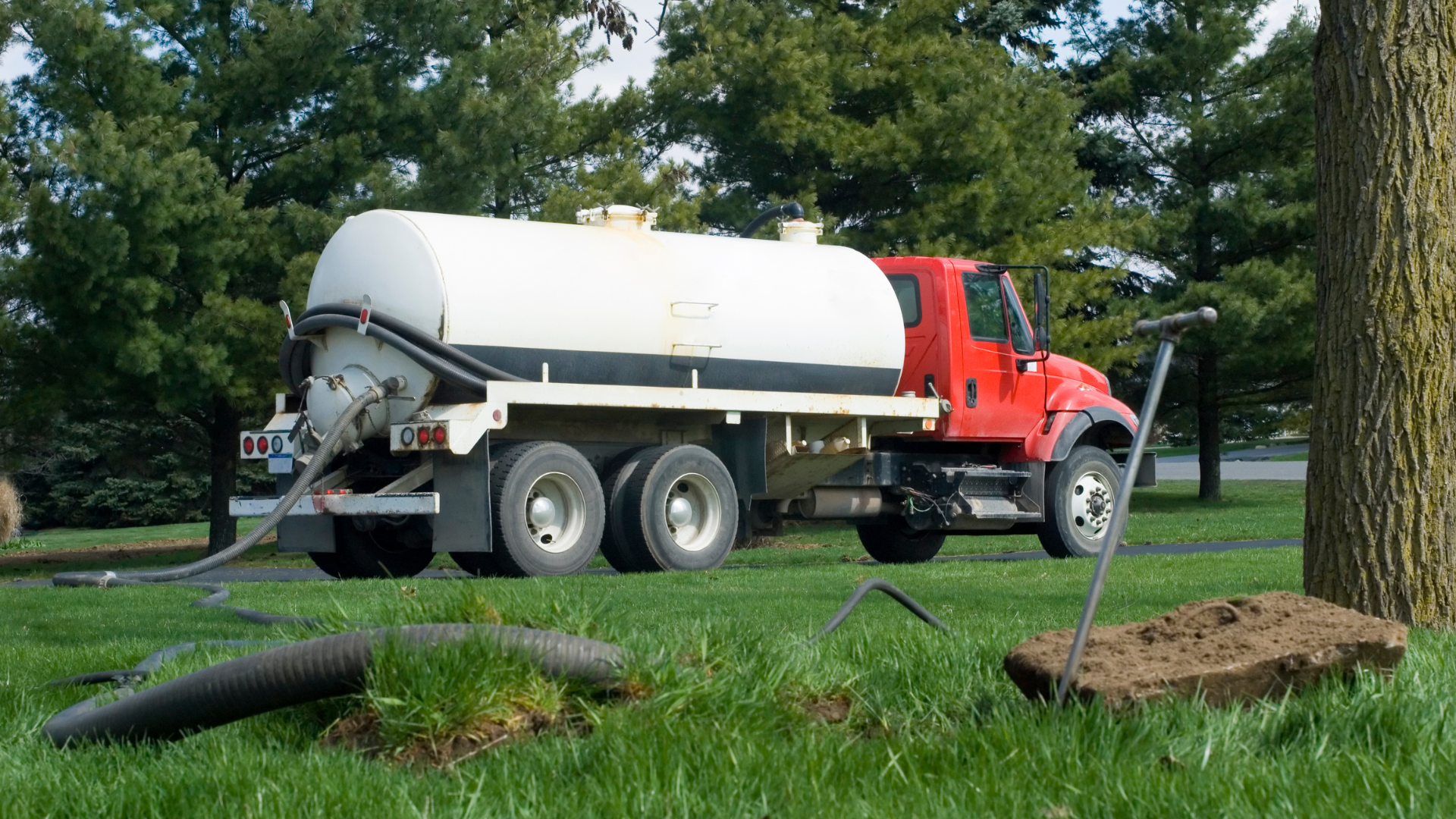 A red septic tank truck on green grass, parked near a tree, with hose in use.