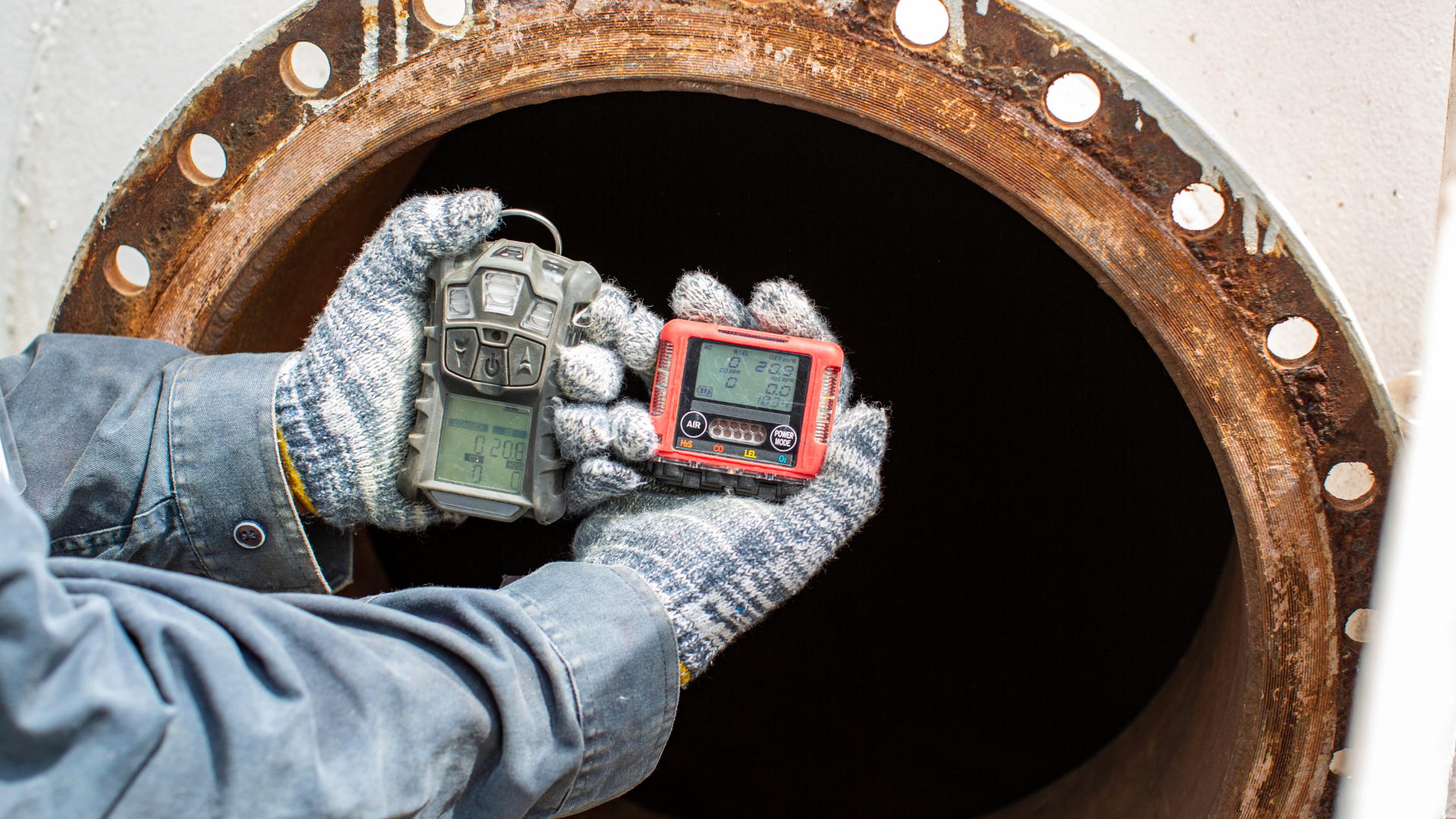 Person holding two gas detectors in front of a large pipe opening, wearing gloves.