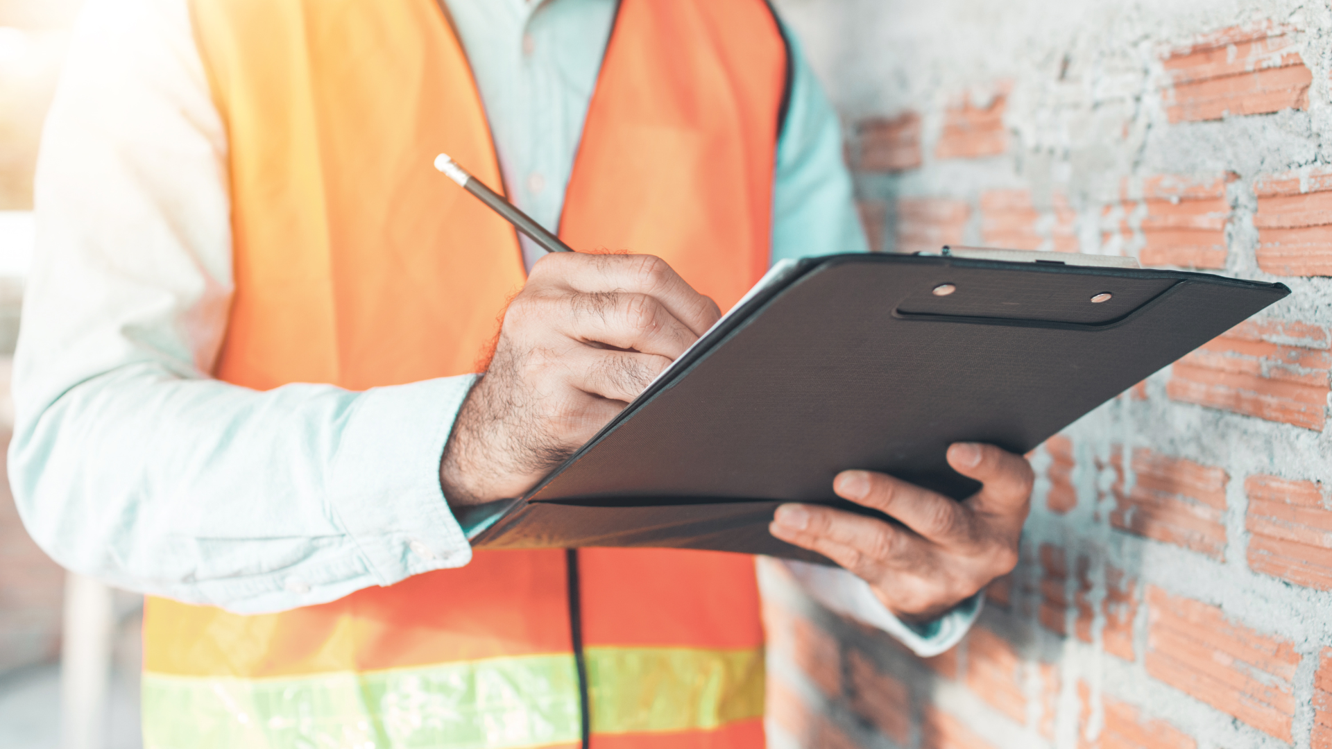Construction worker in orange vest, writing on a clipboard near a brick wall.