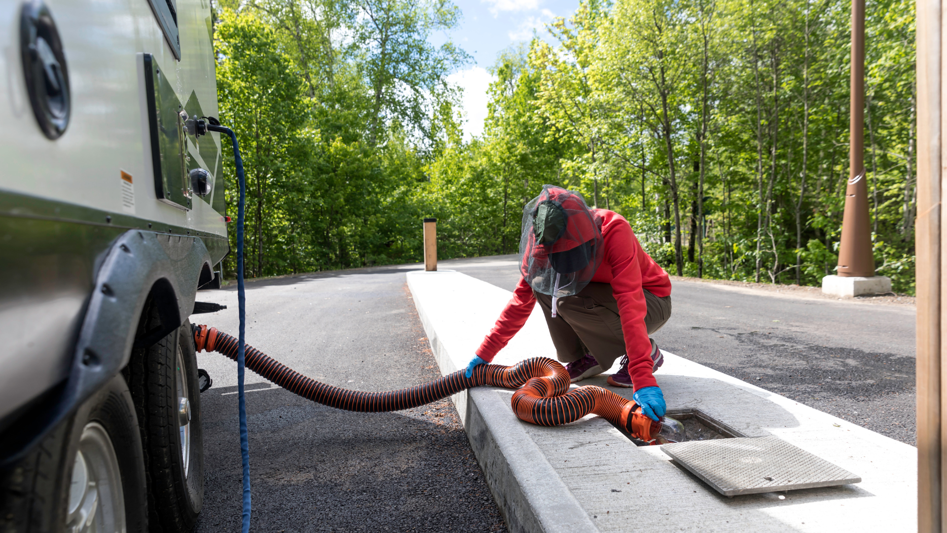 Person emptying RV waste tank. RV parked at campsite with sewer hookup. Person in red jacket.