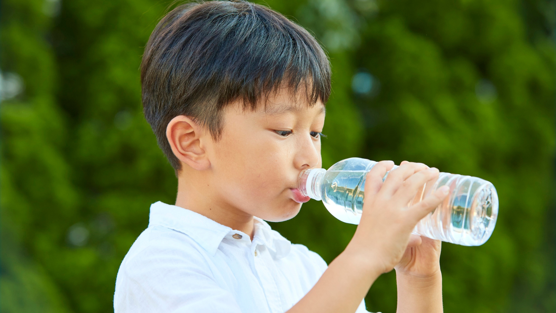 Boy drinking water from a clear plastic bottle, outdoors in front of green foliage.