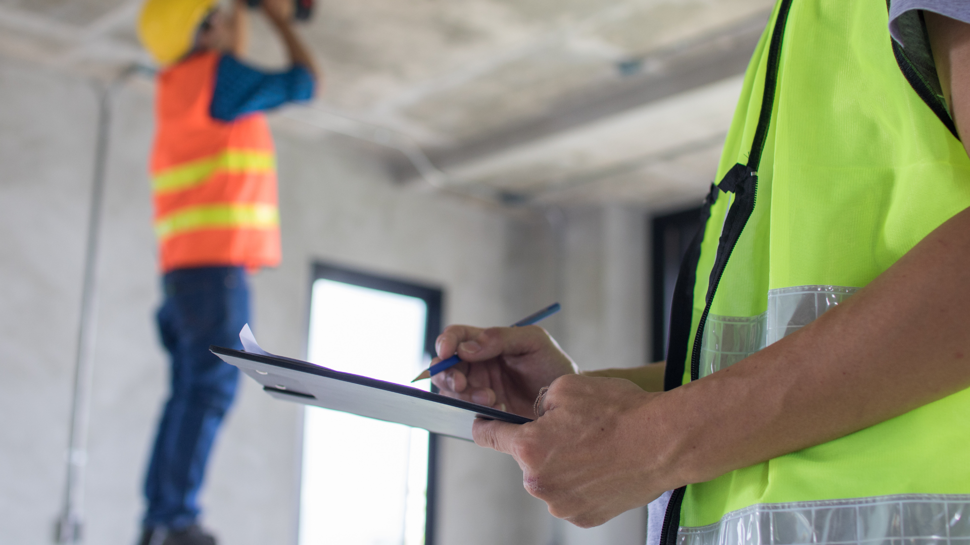 A construction worker in a high-vis vest writes on a clipboard, while another worker in the background checks the ceiling.