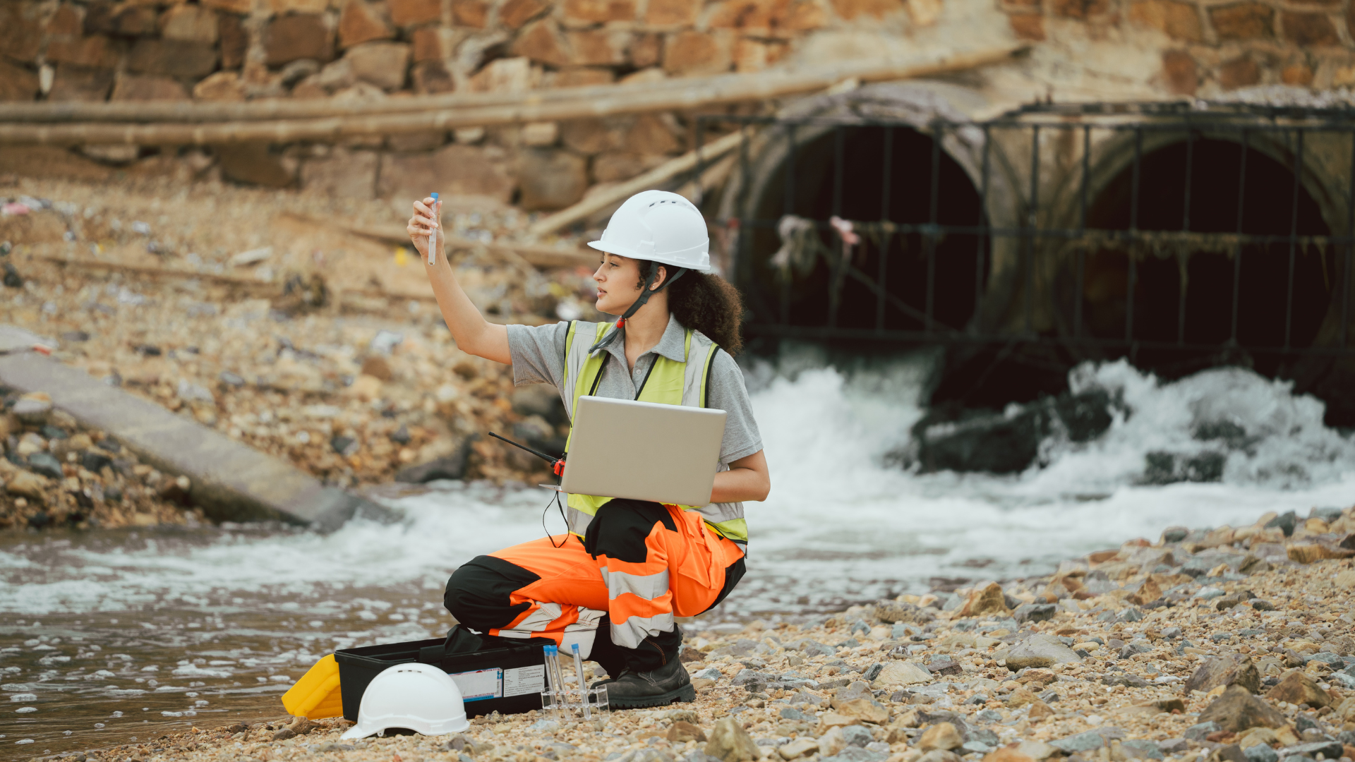 A person in a safety vest and helmet checks water quality near a discharge pipe.