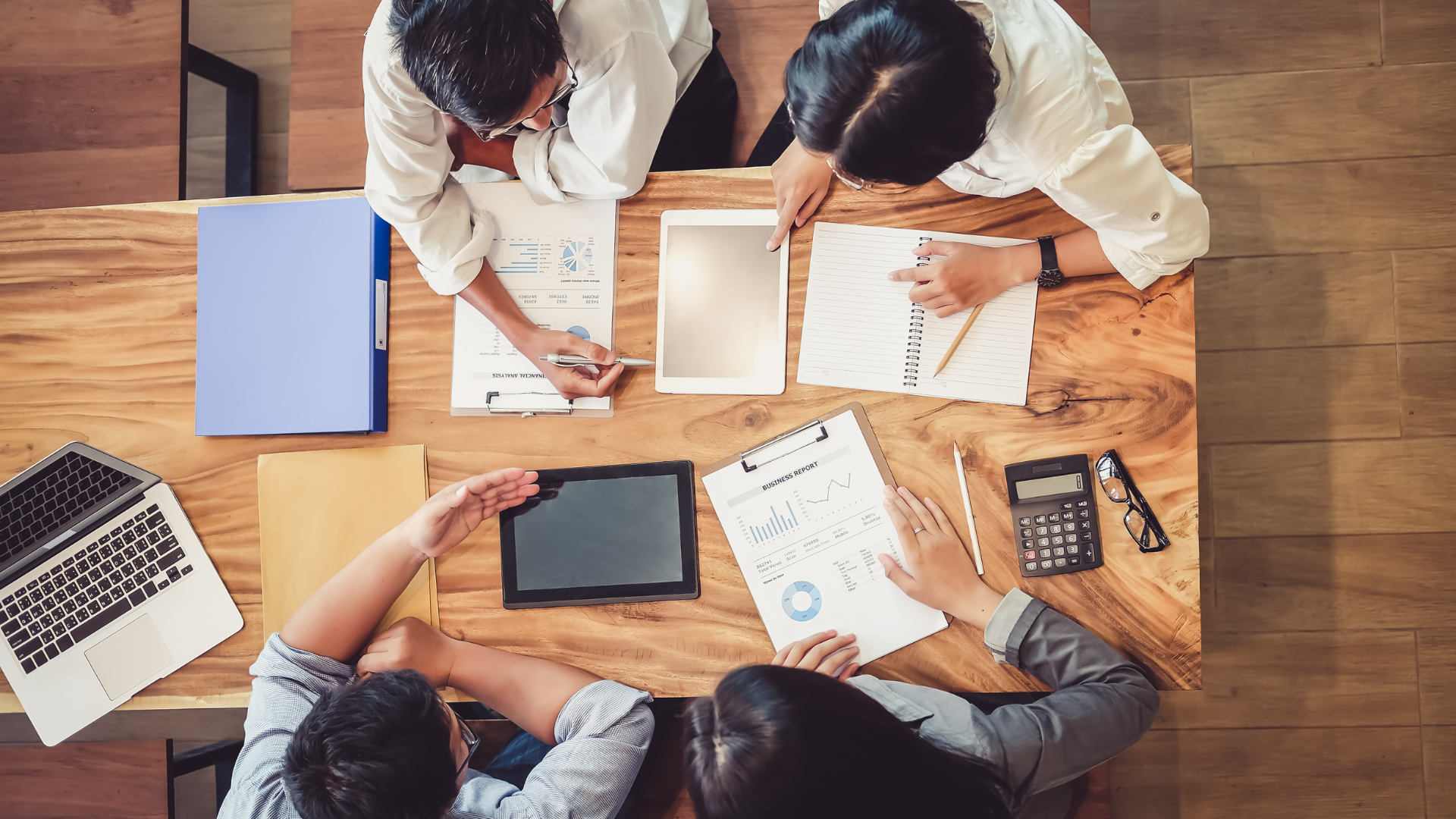 People at a wooden table reviewing documents, using a laptop, tablet, calculator, and pens.