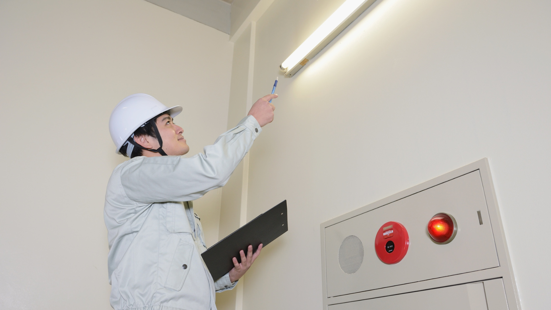 Person in hard hat inspecting a fluorescent light fixture on a white wall, holding a pen and clipboard.