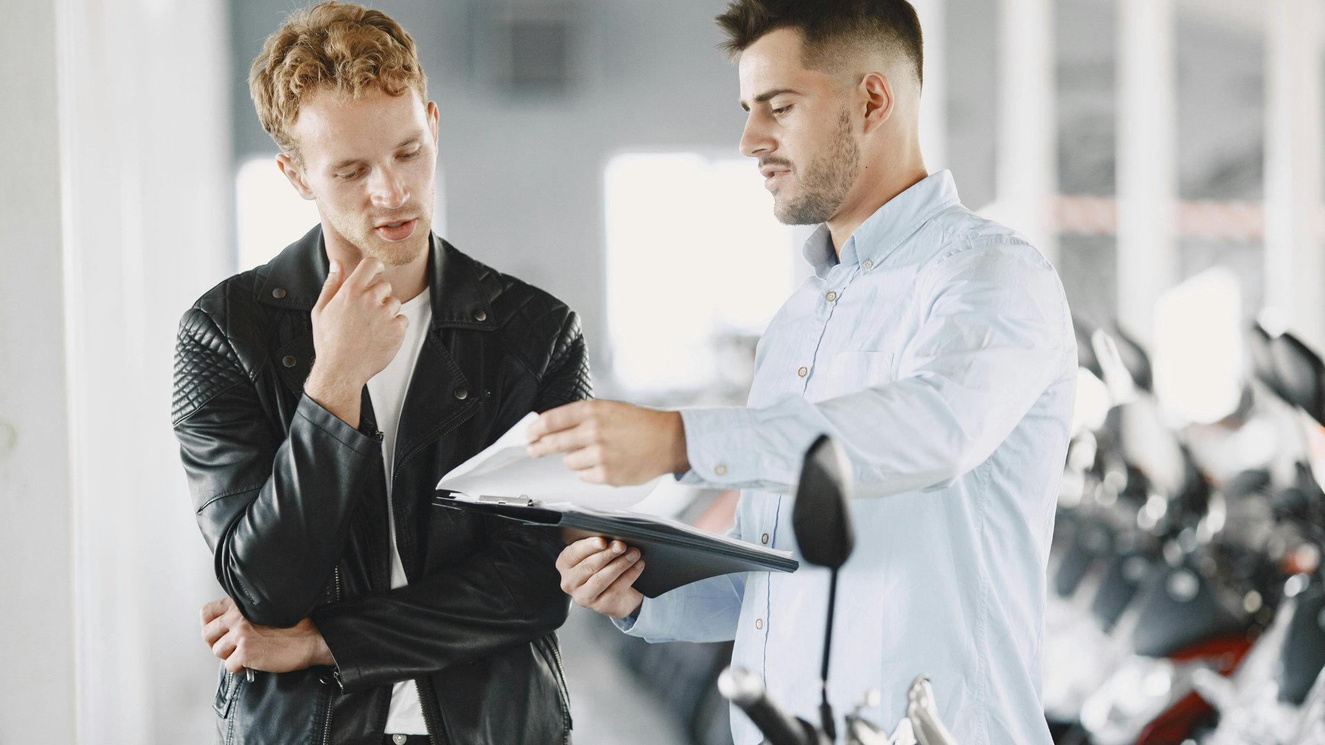 Man in leather jacket ponders documents held by another man in a light blue shirt, in a showroom with motorcycles.