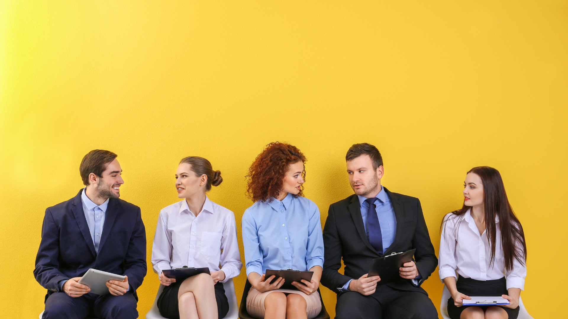 Five people seated in a row, holding documents, against a yellow background. They appear to be in a waiting area.