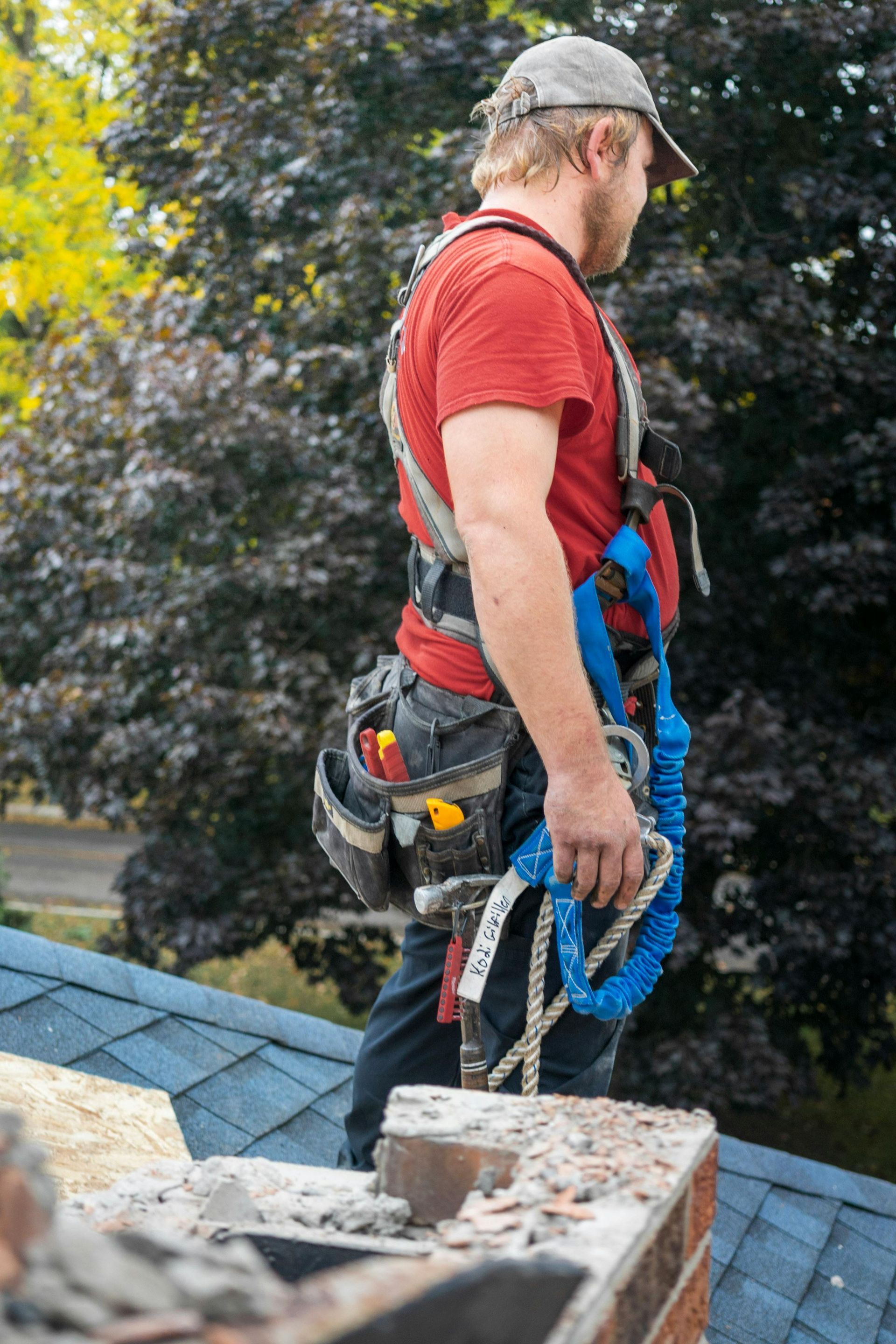 Man on a roof wearing a safety harness, holding a lanyard.  Gray cap, red shirt, tool belt, and blue shingles are visible.